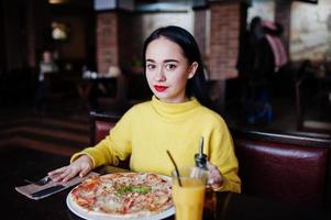 Funny brunette girl in yellow sweater eating pizza at restaurant. photo