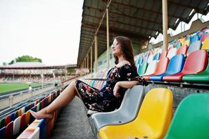 Portrait of a young beutiful girl in dress and sunglasses sitting on the tribunes in stadium. photo