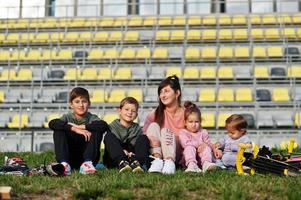 joven madre elegante con cuatro hijos sentados en el césped contra el estadio. la familia deportiva pasa tiempo libre al aire libre con scooters y patines. foto