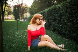 Attractive redhaired woman in eyeglasses, wear on red blouse and jeans skirt posing at green park. photo