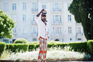 Stylish african american man in white shirt and colored pants with hat and glasses posed outdoor. Black fashionable model boy. photo