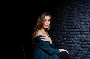 Handsome brunette girl wear on black and green jacket, sitting and posing on chair at studio against dark brick wall. Studio model portrait. photo