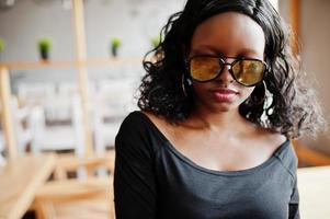 cerrar la cara de una chica afroamericana con gafas de sol posadas en un café moderno. foto