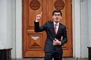 Indian young man at glasses, wear on black suit with red tie posed outdoor against door of building show finger up, he has idea. photo