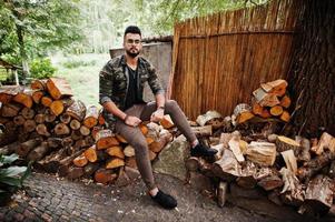 Impresionante y hermoso hombre macho alto de barba árabe con gafas y chaqueta militar posado al aire libre, sentado en tocones. foto