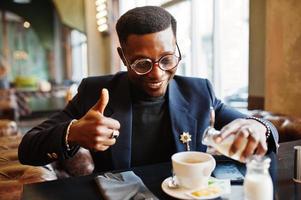 Fashionable african american man in suit and glasses pours cream into coffee at cafe and show thumb up. photo