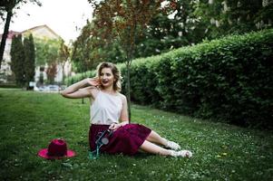 Fashionable and beautiful blonde model girl in stylish red velvet velour skirt, white blouse and hat, sitting on green grass at park with phone and earphones. photo