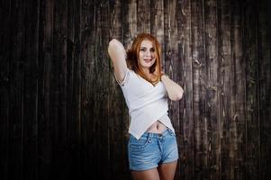 retrato de una hermosa chica pelirroja con camiseta blanca y pantalones cortos de mezclilla posando en el estudio junto a la pared de madera. foto