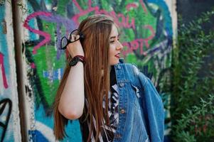 chica hipster casual con estilo en ropa de jeans y gafas contra una gran pared de graffiti. foto