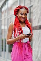 Fashionable african american girl at pink pants and red dreads posed outdoor with mobile phone. photo