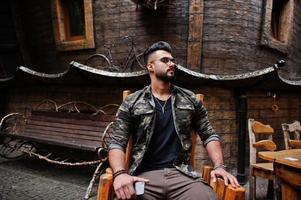 Awesome beautiful tall ararbian beard macho man in glasses and military jacket posed outdoor, sitting on chair against wooden house. photo