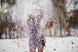 mujer afroamericana de pelo rizado vestida con abrigo de piel de oveja y guantes posados en el día de invierno arroja nieve, enfoque borroso. foto