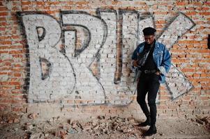 hombre afroamericano con chaqueta de jeans, boina y anteojos contra la pared de graffiti con letrero bruk. foto