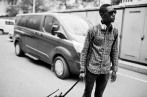 African american man in checkered shirt, sunglasses and earphones with suitcase and backpack. Black man traveler against van car. photo