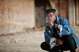 African american man in jeans jacket, beret and eyeglasses, smoking cigar at abandoned factory. photo