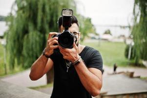Impresionante y hermoso fotógrafo de hombre macho con barba árabe alta con gafas y camiseta negra con cámara profesional en las manos. foto