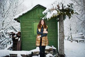 Elegance curly girl in fur coat at snowy forest park agasinst green wooden house at winter. photo