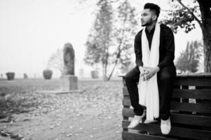 Indian stylish man in black traditional clothes with white scarf sitting outdoor. photo