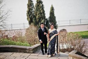 Elegant and fashionable indian friends couple of woman in saree and man in suit posed on stairs against lake. photo