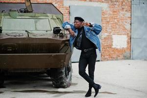 African american man in jeans jacket, beret and eyeglasses, with cigar posed against btr military armored vehicle. photo