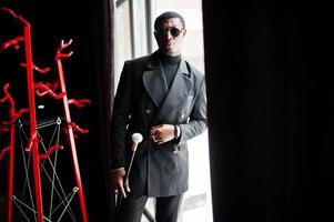 Stylish african american gentleman in elegant black jacket and sunglasses, holding retro walking stick as cane flask or tippling cane with golden diamond ball handle. Rich fashionable afro man. photo
