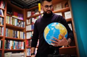 Tall smart arab student man, wear on violet turtleneck and eyeglasses, at library holding earth globe at hands. photo