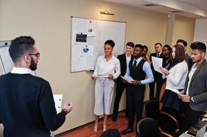 Mixed race business coach presenting report standing near whiteboard pointing on sales statistic shown on diagram and chart teach diverse company members gathered together in conference room. photo