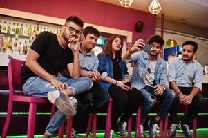 Group of stylish asian friends wear on jeans sitting at chairs against bar in club and makes selfie together. photo