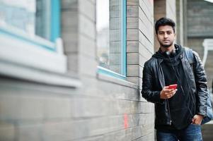 hombre asiático elegante e informal con chaqueta de cuero negro, auriculares con teléfono móvil rojo en las manos posado en la calle. foto