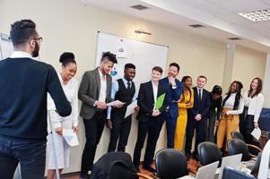 Mixed race business coach presenting report standing near whiteboard pointing on sales statistic shown on diagram and chart teach diverse company members gathered together in conference room. photo