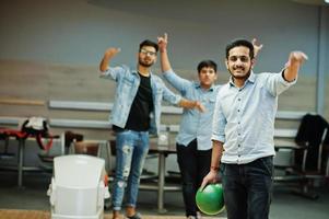 South asian man in jeans shirt standing at bowling alley with ball on hands. Guy is preparing for a throw. Friends support him loudly. photo