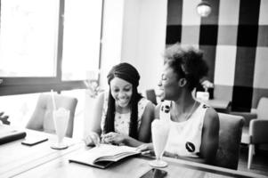 Two black african girlfriends at summer dresses drinking milkshake cocktails and looking in menu at cafe. photo