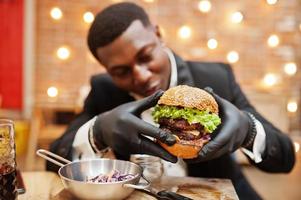 Respectable young african american man in black suit and gloves for food sitting in restaurant with tasty double burger and soda drink. photo