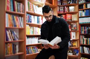 estudiante árabe alto e inteligente, vestido con chaqueta negra de jeans y anteojos, en la biblioteca con un libro en las manos. foto