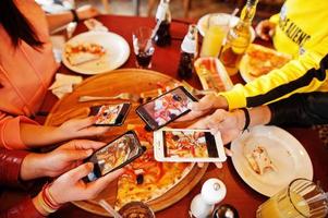 Hands of friends making photo by phones of pizza during party at pizzeria. Happy people having fun together.