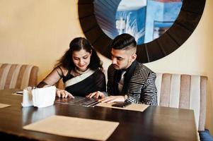 Lovely indian couple in love, wear at saree and elegant suit, posed on restaurant. They looking at menu to choose a dish. photo