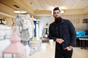 Arab man wear on black jeans jacket and eyeglasses in cafe drink coffee at bar. Stylish and fashionable arabian model guy. photo