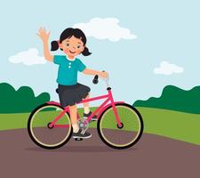 niña feliz montando en bicicleta divirtiéndose en el parque en un día soleado agitando la mano vector