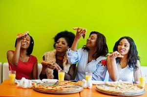 Four young african girls in bright colored restaurant eating pizza slices in hands. photo