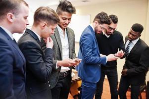 seis hombres de negocios multirraciales parados en la oficina y usan teléfonos móviles. grupo diverso de empleados varones en ropa formal con teléfonos celulares. foto