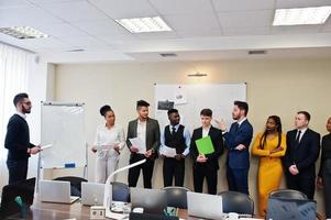 Mixed race business coach presenting report standing near whiteboard pointing on sales statistic shown on diagram and chart teach diverse company members gathered together in conference room. photo