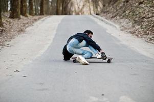 fallar al caer de una patineta. hombre árabe de estilo callejero en anteojos con longboard longboarding por el camino. foto