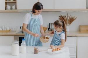 Happy mother and daughter bake together in kitchen, use different ingredients, wear aprons, stand against kitchen interior, girl pours milk in bow. Caring mommy teaches child to cook or make dough photo