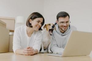 Photo of smiling woman and man work on modern laptop computer, dog yawns, buy furniture for new apartment, lie on floor in spacious light room, have glad expressions. Housewarming and repair concept