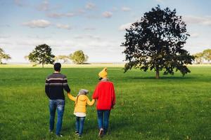 la familia feliz se toma de la mano, camina por el prado o el campo verde, retrocede ante la cámara, disfruta de la libertad, la unión y los hermosos paisajes. pasar tiempo en familia juntos. concepto de relajación y ocio foto