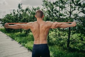 la vista posterior del deportista irreconocible estira la banda de resistencia elástica, demuestra un cuerpo musculoso fuerte, usa equipo deportivo, se para afuera, disfruta de los ejercicios deportivos. concepto de culturismo foto