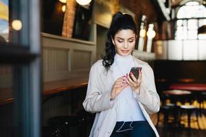 Pretty brunette woman with pony tail and appealing appearance sitting in cafe using her smartphone surfing internet using free wifi conncection and listening to her favourite music with earphones photo