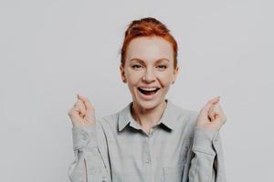 feliz mujer pelirroja emocionada sosteniendo los puños apretados y celebrando el éxito en el estudio foto