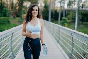 Active beautiful sporty woman in active wear holds mobile phone, listens music in earphones, takes break after jogging holds bottle of fresh water poses at bridge outdoors. Fitness, lifestyle concept photo