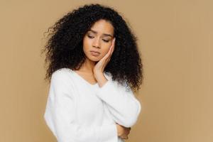 Melancholic sad lonely woman focused down with thoughtful expression, keeps hand on cheek, has calm expression, wears white sweater, has curly hair, isolated on brown studio wall. Depression photo